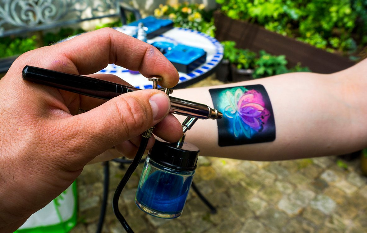 Malowanie aerografem wzoru kwiatu na ramieniu, widoczny aerograf, farba i fragment otoczenia. Detaliczny widok na proces tworzenia tymczasowego tatuażu.
