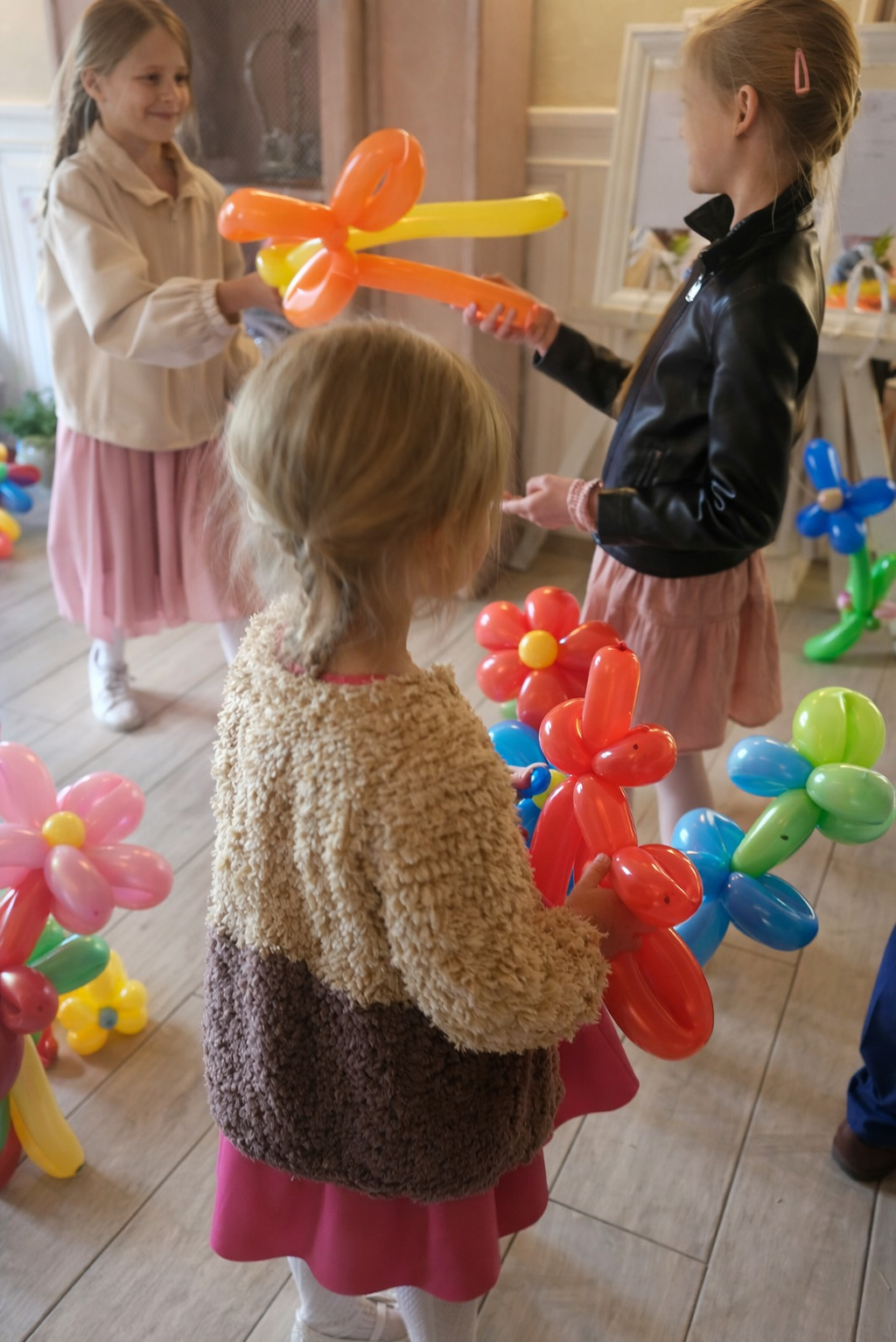 Dziewczynki bawią się balonami w kształcie kwiatów i zwierząt podczas imprezy urodzinowej. Jasne wnętrze, drewniana podłoga, dekoracje balonowe.