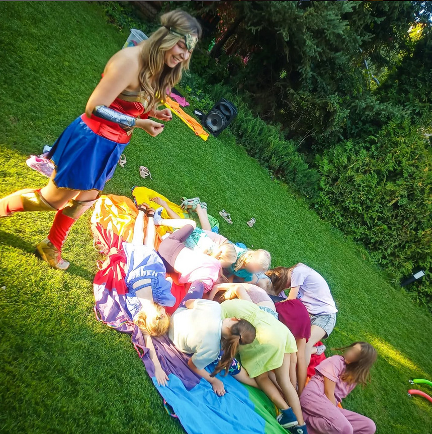 Animator w stroju Wonder Woman prowadzi zabawę z grupą dzieci leżących na kolorowej chuście na trawie podczas imprezy plenerowej. Widoczny głośnik i zieleń w tle.