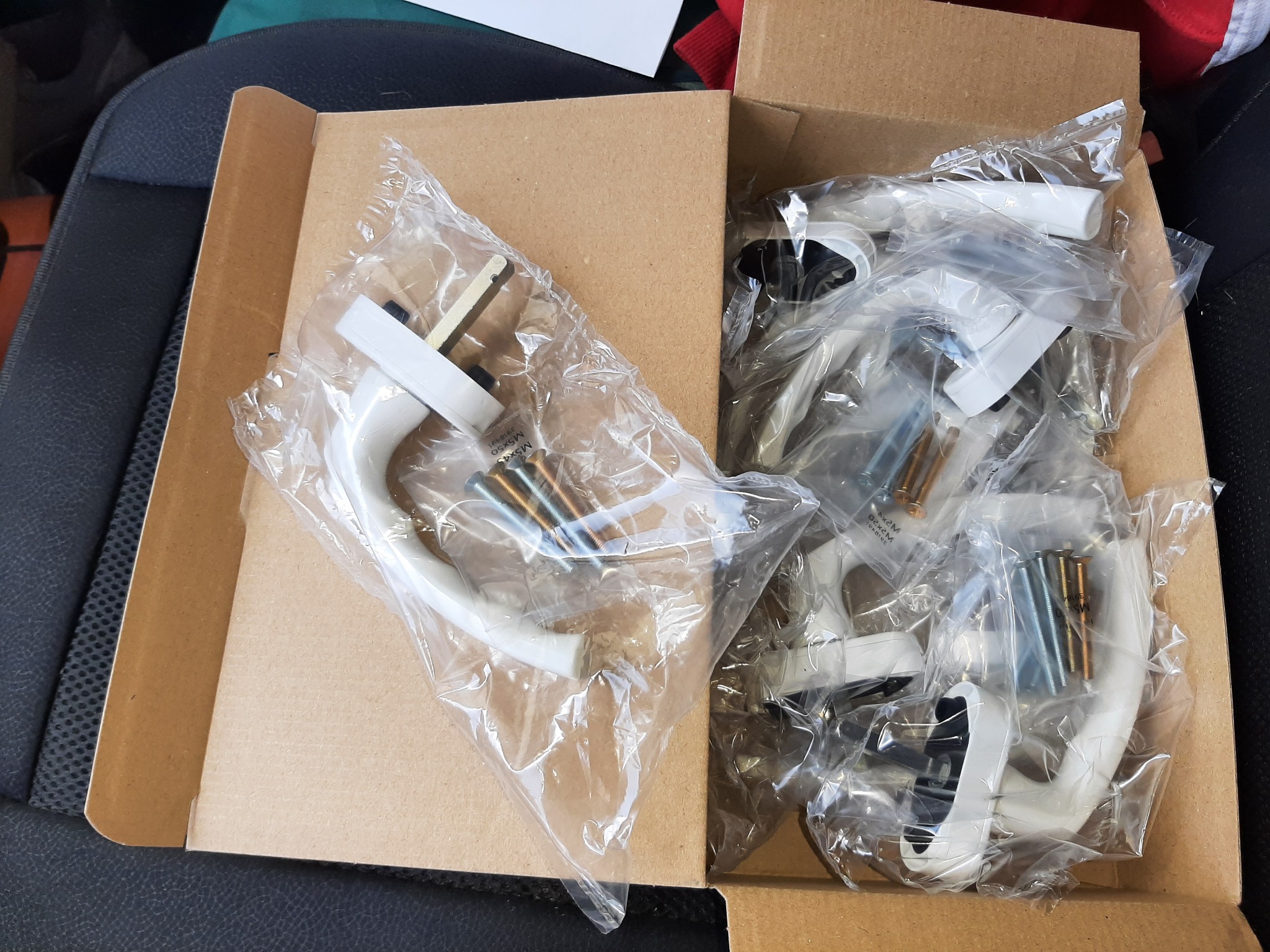 Nowe, białe klamki do drzwi zapakowane w folię i umieszczone w kartonowym pudełku, widoczne śruby montażowe.