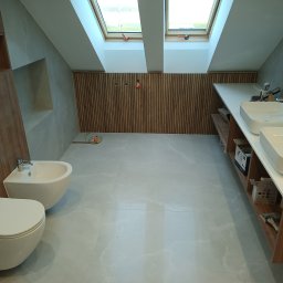 ArrowBud - Wnętrze łazienki na poddaszu z dwoma oknami dachowymi, wykończone płytkami w odcieniach szarości, z drewnianą boazerią na ścianie, białą ceramiką sanitarną i elementami wyposażenia w trakcie montażu.