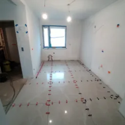 Wnętrze w trakcie układania jasnych, marmurkowych płytek podłogowych z użyciem systemu poziomowania, widoczne krzyżyki dystansowe i poziomica przy ścianie.