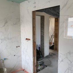 ArrowBud - Wnętrze łazienki w trakcie układania płytek imitujących marmur, widoczne niedokończone ściany, otwory na instalacje, narzędzia i odkurzacz przemysłowy.