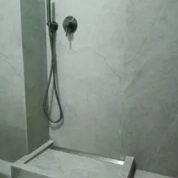 Szara kabina prysznicowa z odpływem liniowym, wykończona płytkami imitującymi kamień, z zamontowaną czarną baterią i słuchawką prysznicową.