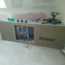 ArrowBud - Szara obudowa z płyt budowlanych Ultrament pod wannę w trakcie montażu, widoczne przyłącza wodne, narzędzia i akcesoria na górze obudowy, ściany w surowym stanie z widocznymi notatkami.