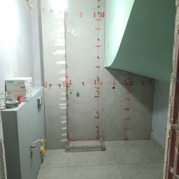 ArrowBud - Świeżo wykafelkowana ściana w łazience z użyciem poziomowania płytek, widoczne pudełka z armaturą JOMO i fragment nieotynkowanej ściany.