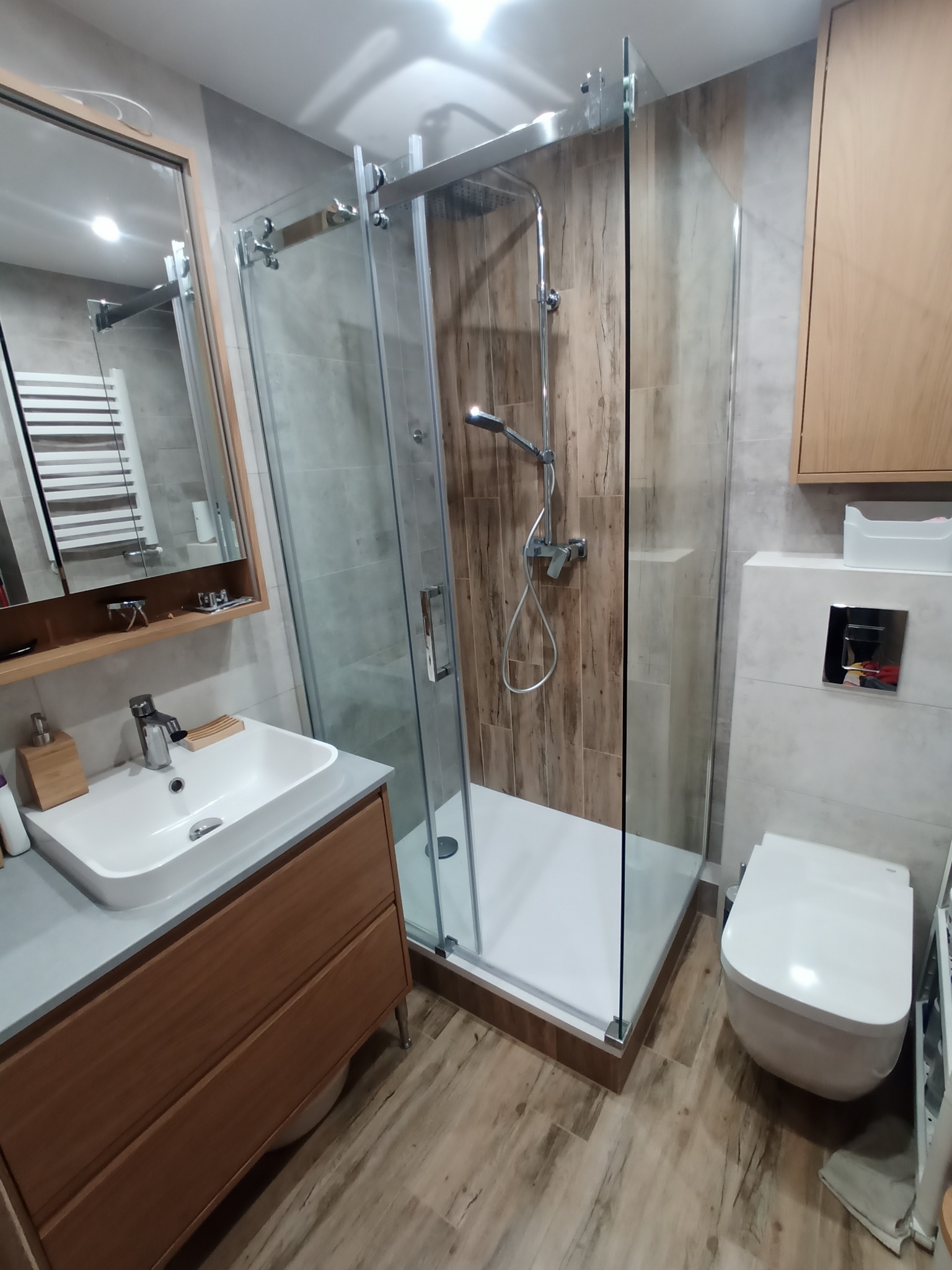 Nowoczesna łazienka z kabiną prysznicową ze szklanymi drzwiami przesuwnymi, wiszącą toaletą z chromowanym przyciskiem spłukiwania i umywalką nablatową na drewnianej szafce, całość w odcieniach...