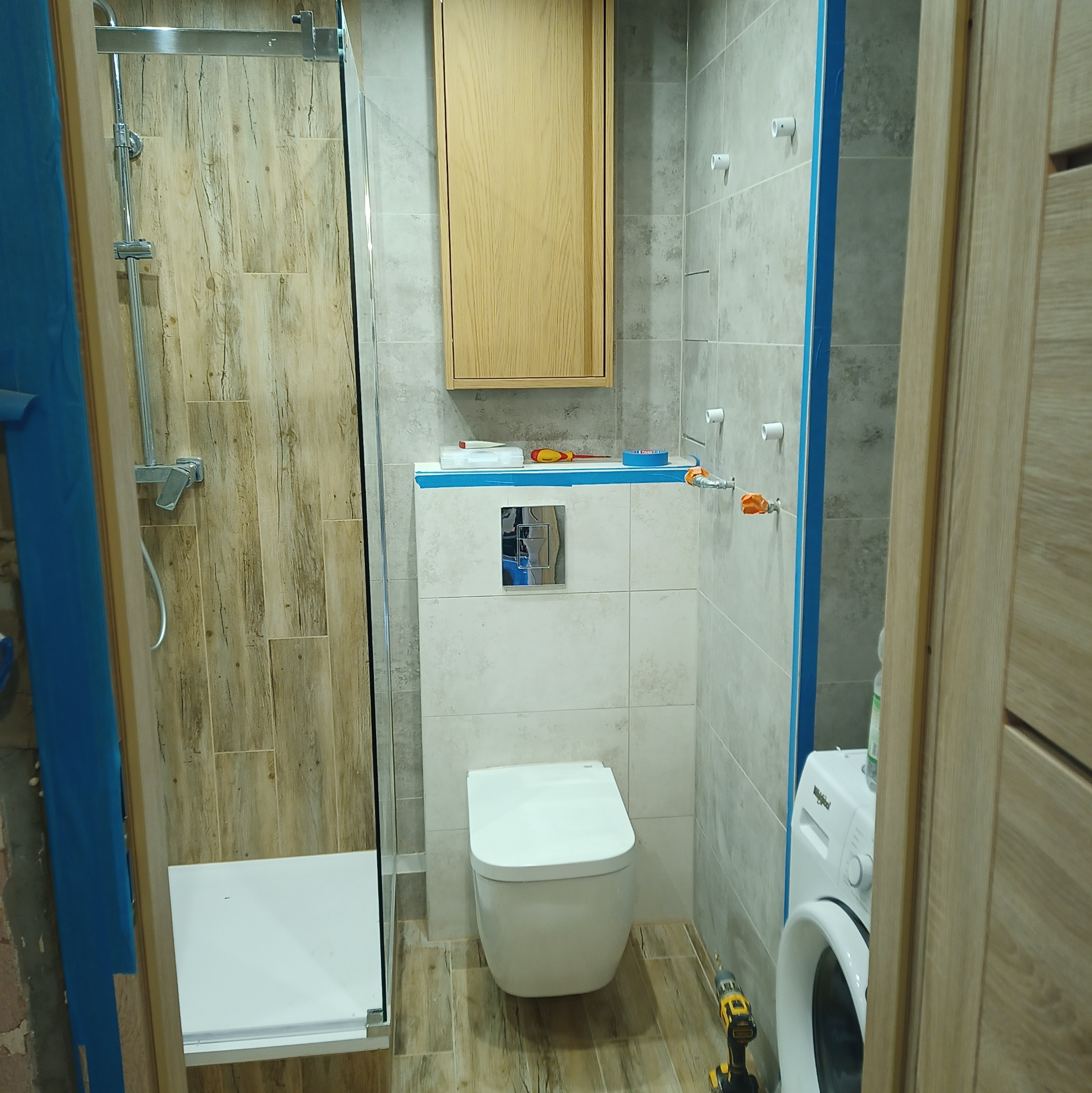 Wnętrze łazienki w trakcie prac, widoczna kabina prysznicowa z drewnopodobnymi płytkami, podwieszana toaleta, pralka i szafka wisząca, zabezpieczone niebieską taśmą malarską.