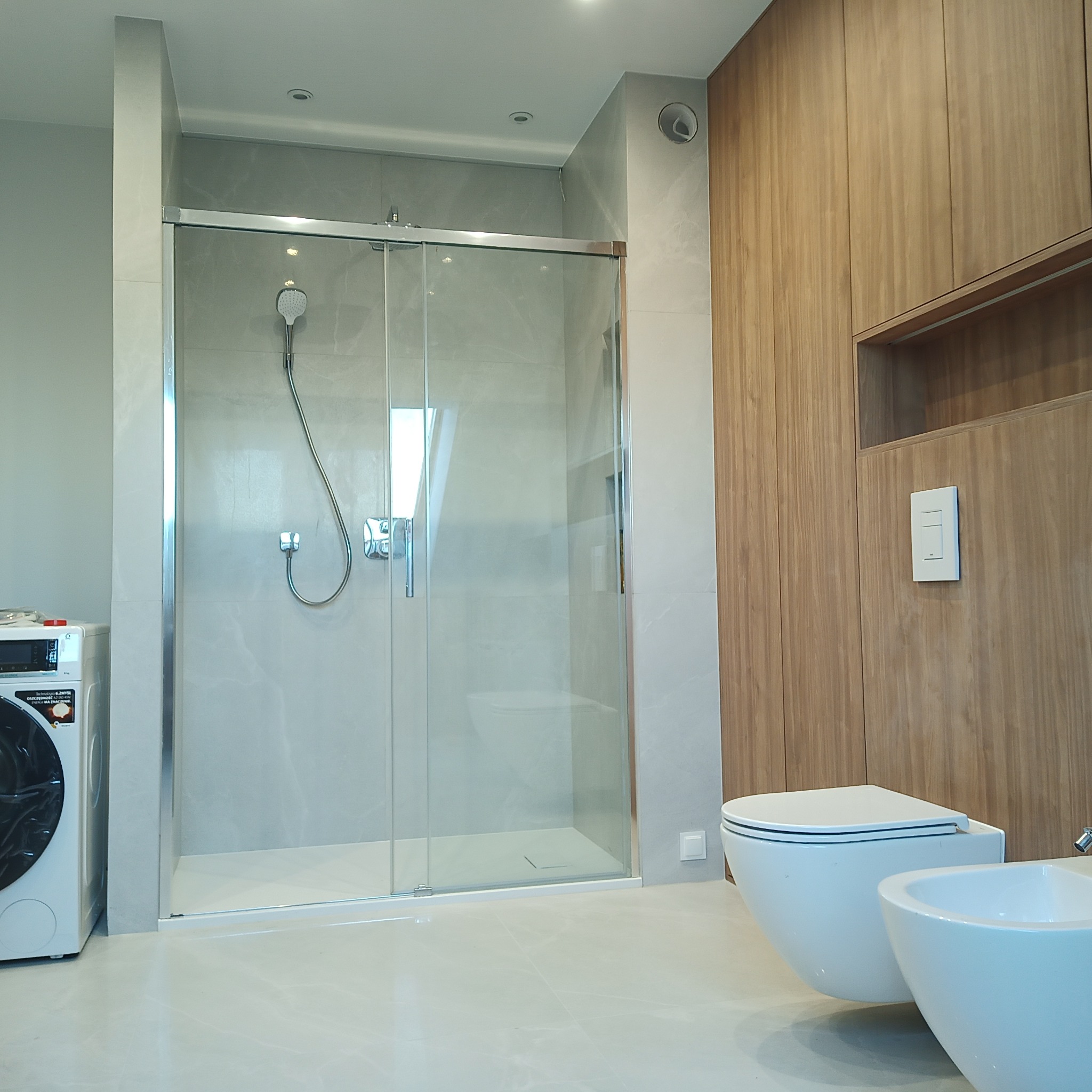 Nowoczesna łazienka z kabiną prysznicową typu walk-in z przesuwnymi drzwiami, podwieszaną toaletą i bidetem, pralką oraz drewnianą zabudową ściany z wnęką.