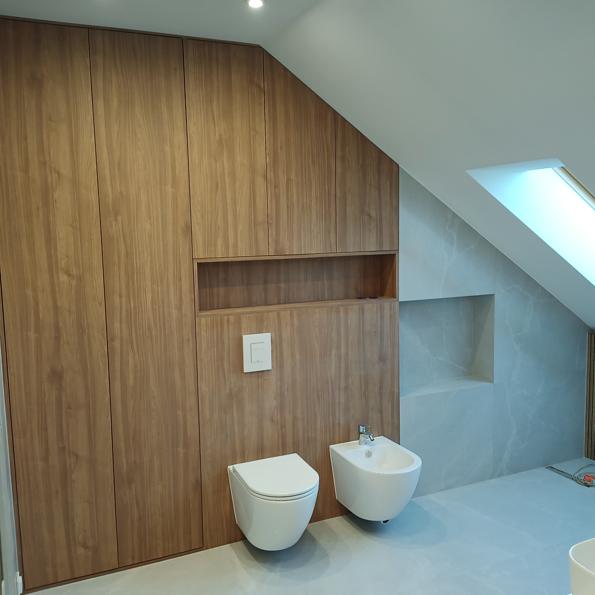 Nowoczesna łazienka na poddaszu ze skośnym sufitem, drewnianą zabudową meblową z wnęką i białym osprzętem sanitarnym: toaleta podwieszana i bidet.