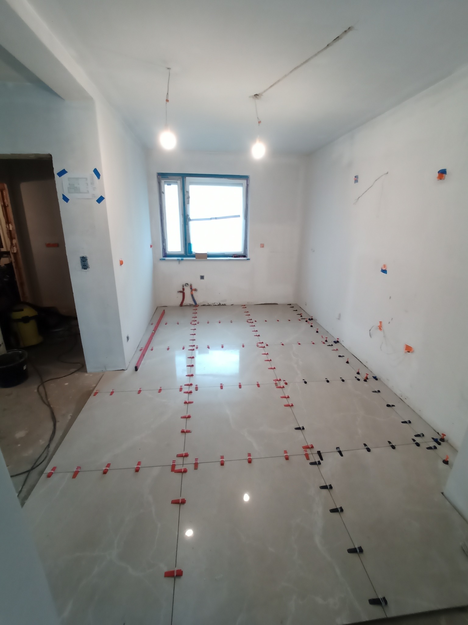 Wnętrze w trakcie układania jasnych, marmurkowych płytek podłogowych z użyciem systemu poziomowania, widoczne krzyżyki dystansowe i poziomica przy ścianie.