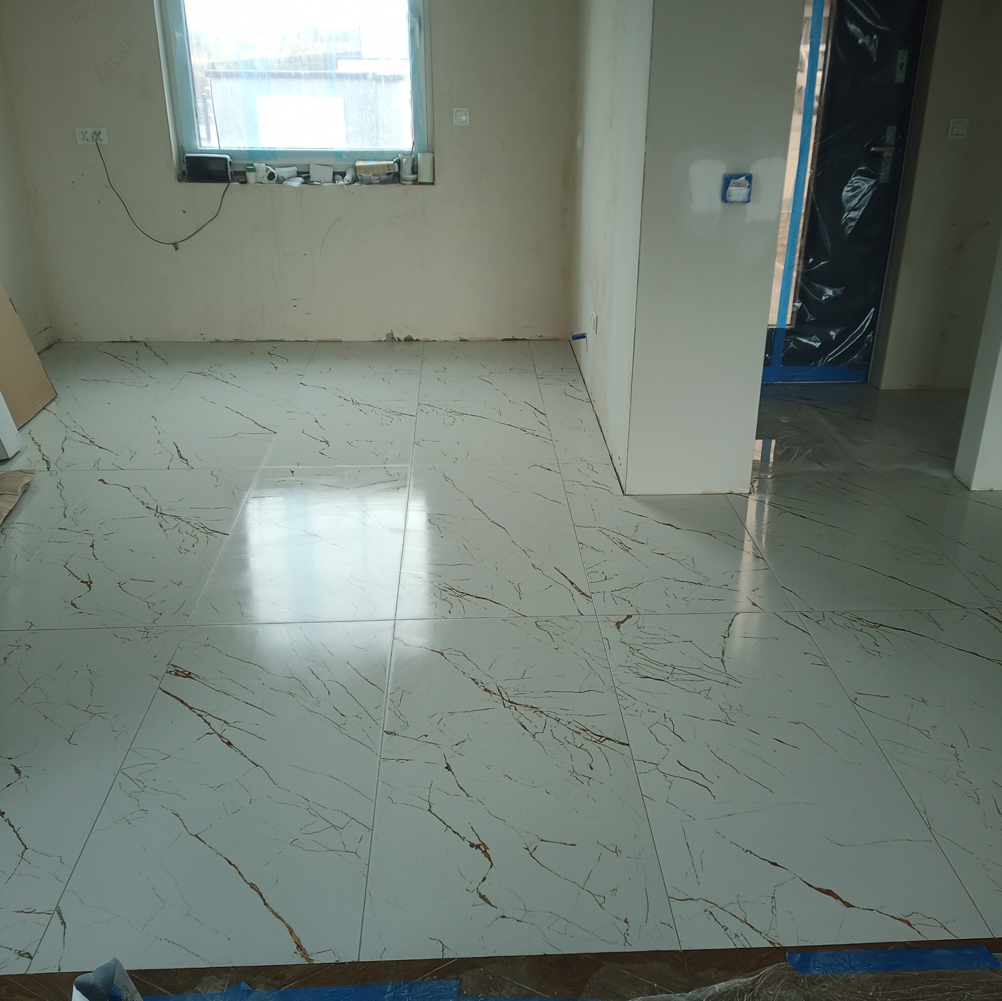 Wyłożona jasnymi płytkami podłoga z wzorem marmuru w trakcie remontu, widoczna folia ochronna na drzwiach i niedokończone ściany.