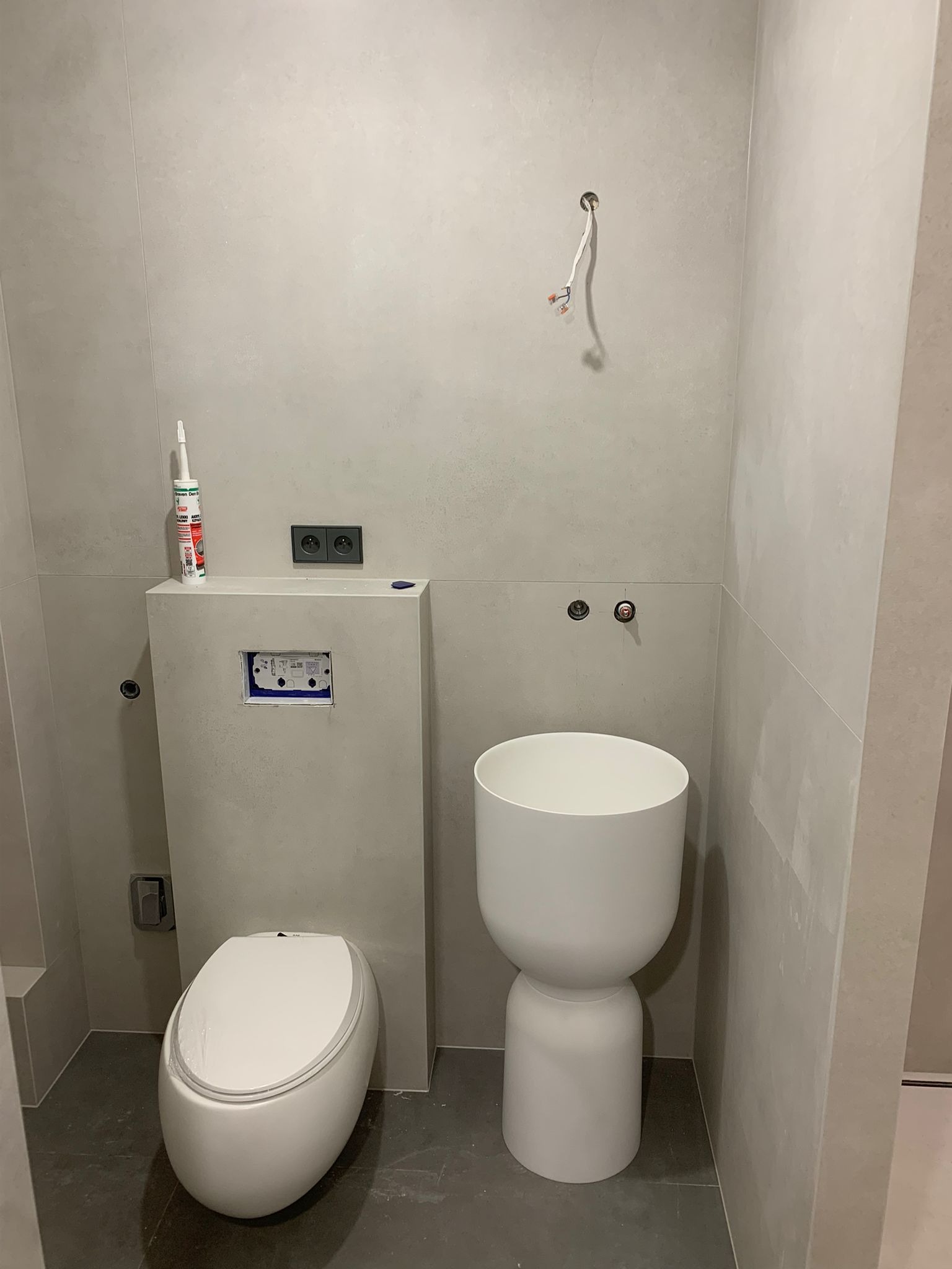 Zawieszona miska WC i umywalka wolnostojąca w trakcie montażu w łazience wykończonej szarymi płytkami, widoczne wystające przewody elektryczne i niedokończone fugi.