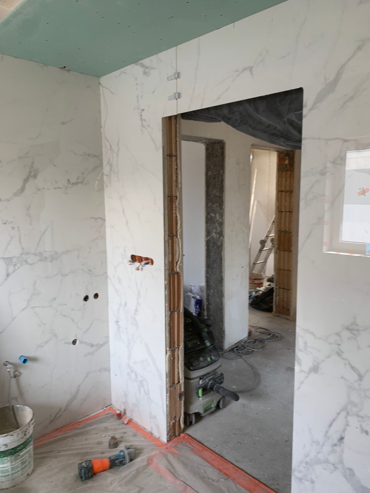 Wnętrze łazienki w trakcie układania płytek imitujących marmur, widoczne niedokończone ściany, otwory na instalacje, narzędzia i odkurzacz przemysłowy.