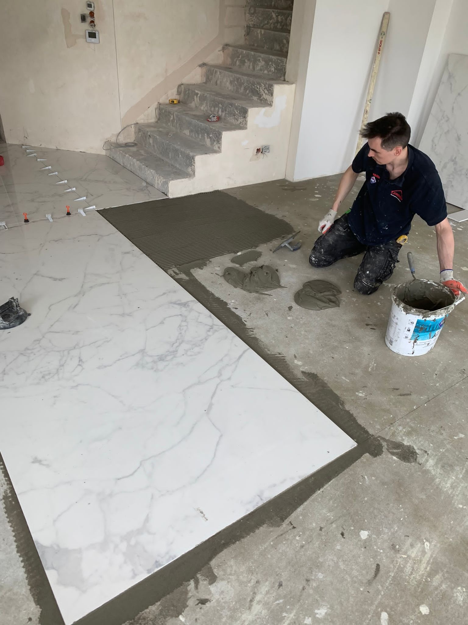 Układanie wielkoformatowych płytek imitujących marmur na podłodze w pomieszczeniu ze schodami; widoczny glazurnik nanoszący klej, poziomica oparta o ścianę i krzyżyki dystansowe.