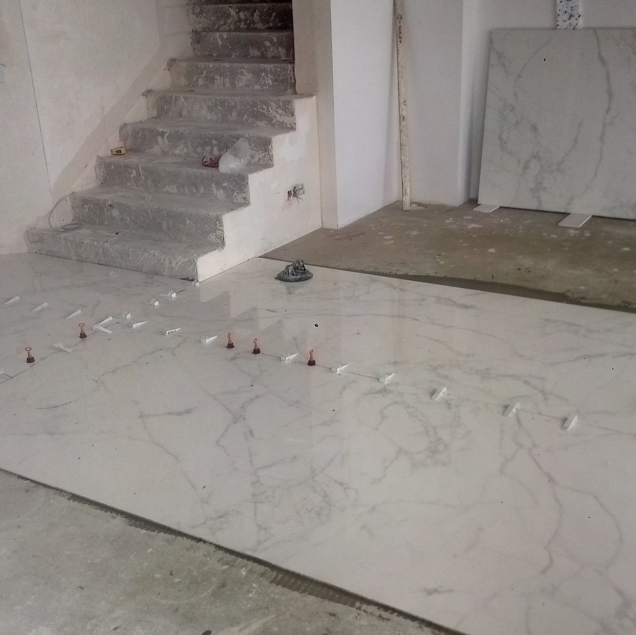 Układanie białych marmurowych płytek podłogowych z użyciem poziomujących klipsów i klinów w pomieszczeniu z surowymi schodami i płytą marmurową opartą o ścianę.