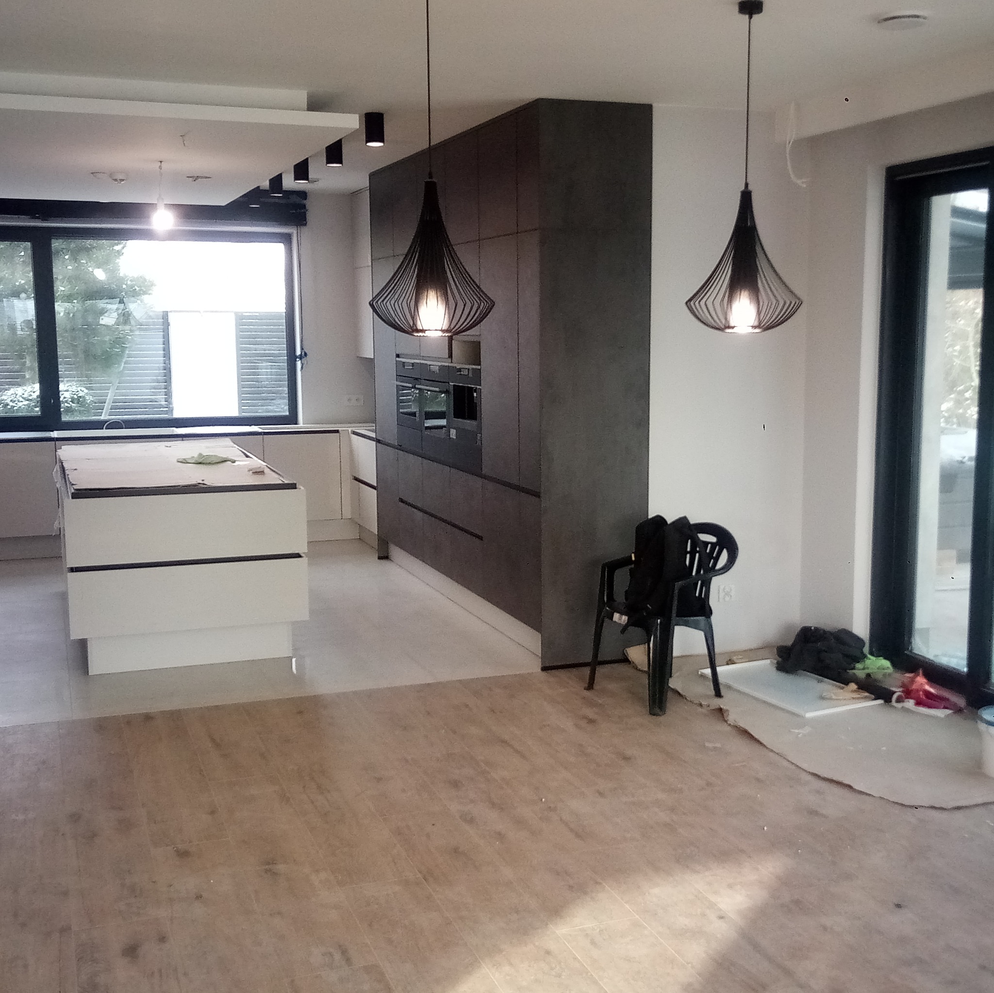 Wnętrze kuchni w trakcie remontu: wyspa kuchenna zabezpieczona kartonem, szare szafki w zabudowie, nowoczesne lampy wiszące z czarnymi kloszami, podłoga częściowo wyłożona panelami, widoczne ślady...