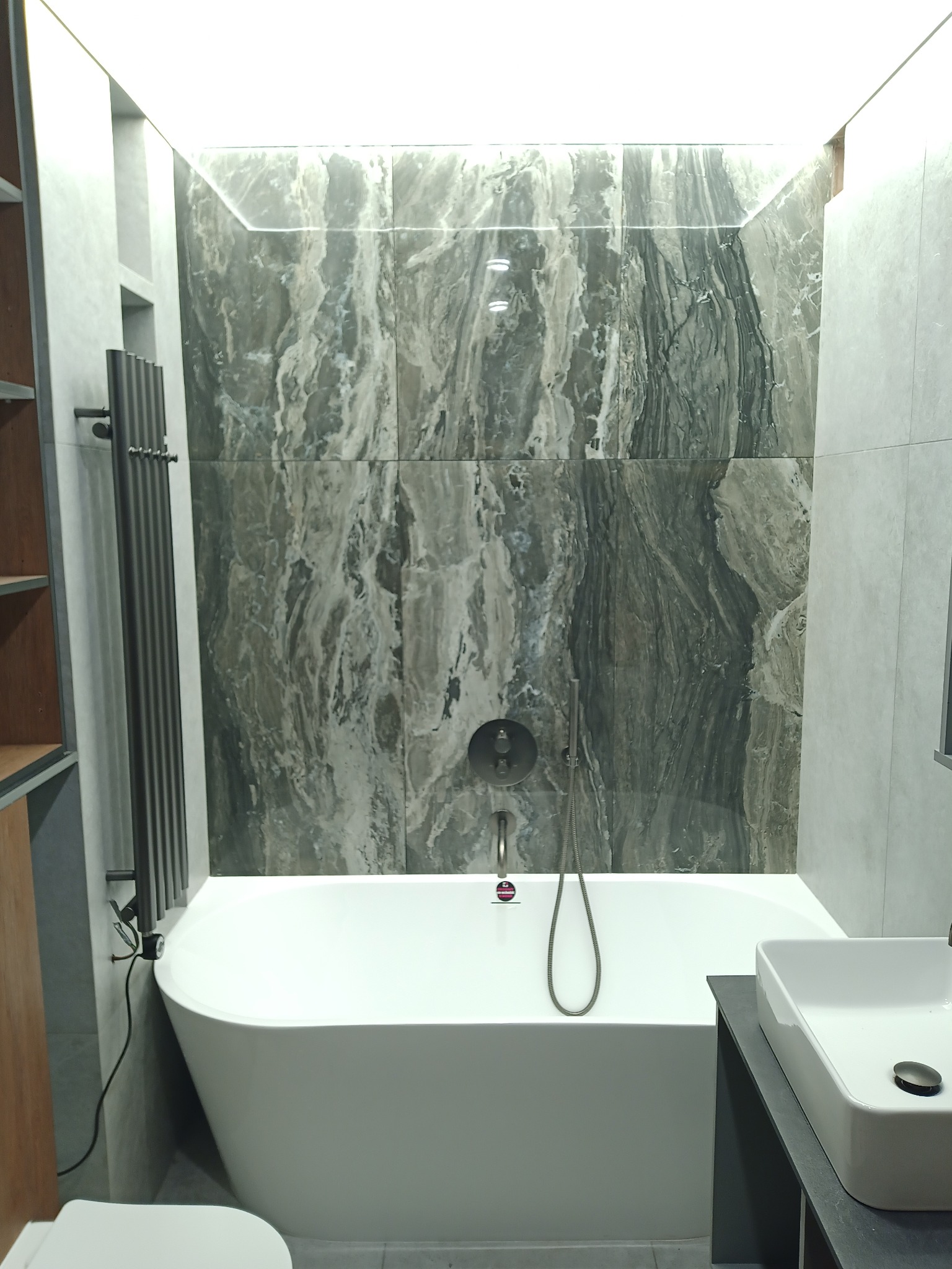 Nowoczesna łazienka z białą wanną wolnostojącą, marmurowe płytki na ścianie podświetlone ledami, grzejnik drabinkowy i umywalka nablatowa.