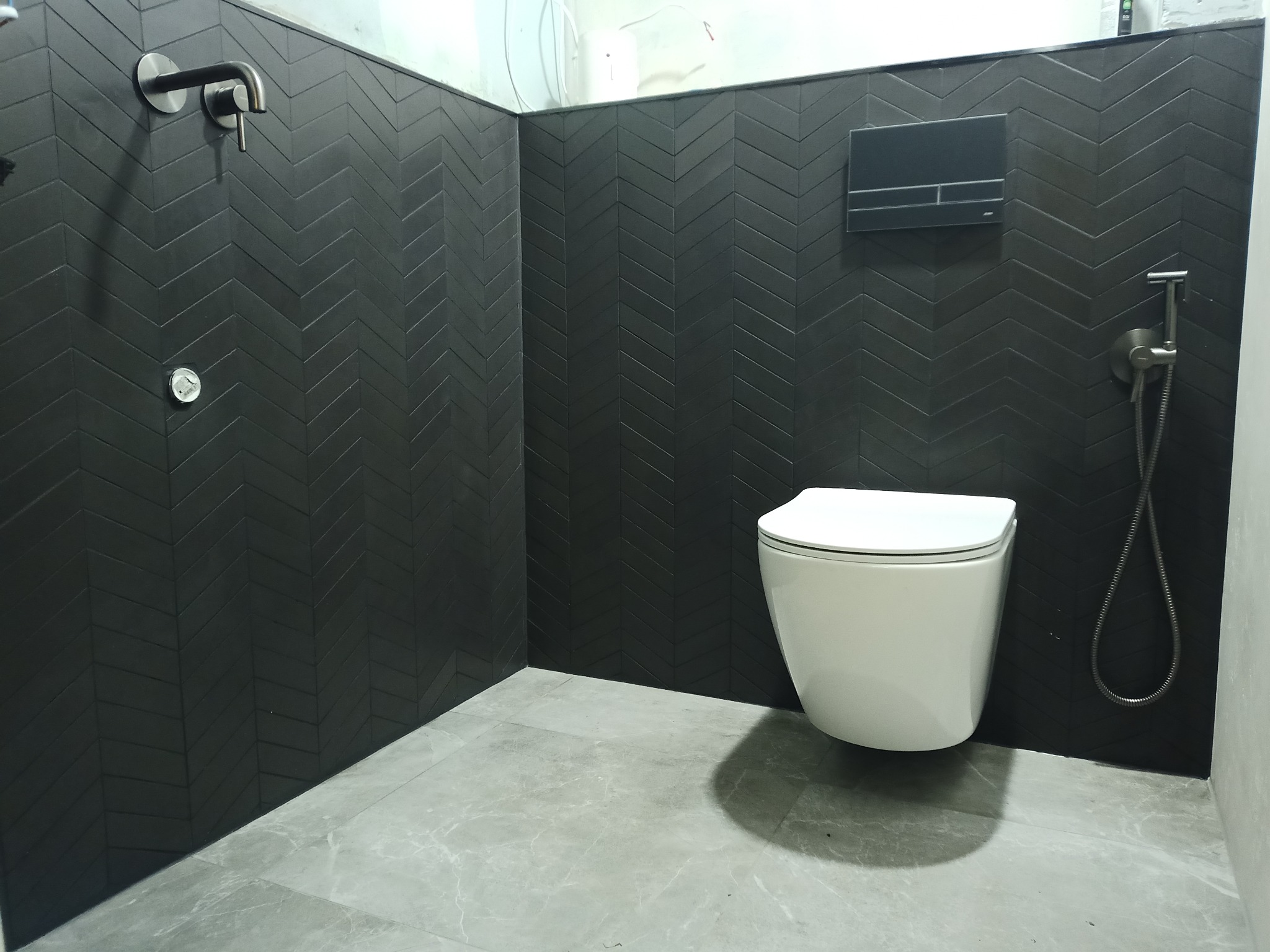 Nowoczesna łazienka z czarnymi płytkami ułożonymi w jodełkę, wisząca toaleta z podtynkową spłuczką, szara marmurowa podłoga i bateria prysznicowa w kolorze grafitowym.