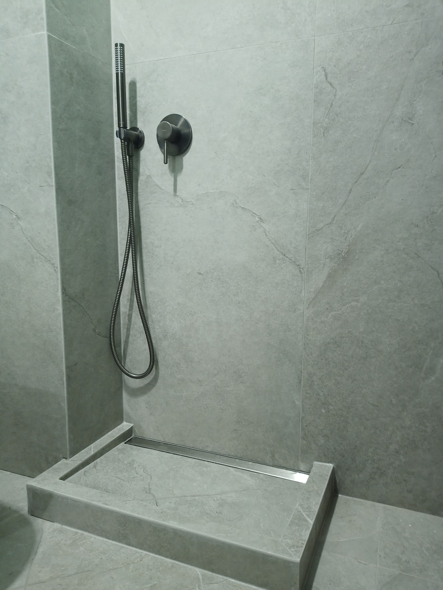 Szara kabina prysznicowa z odpływem liniowym, wykończona płytkami imitującymi kamień, z zamontowaną czarną baterią i słuchawką prysznicową.
