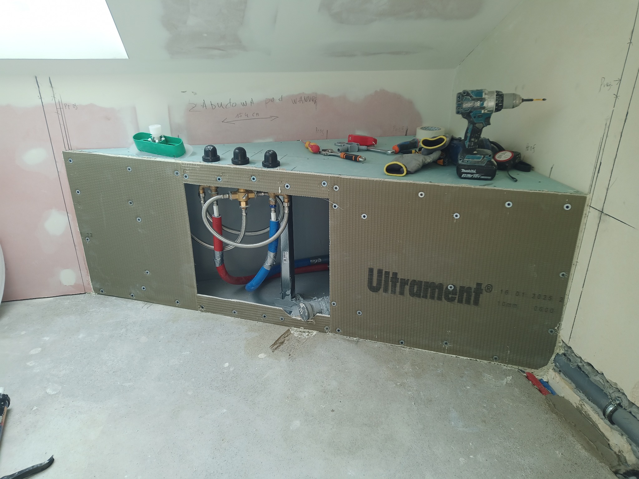 Szara obudowa z płyt budowlanych Ultrament pod wannę w trakcie montażu, widoczne przyłącza wodne, narzędzia i akcesoria na górze obudowy, ściany w surowym stanie z widocznymi notatkami.