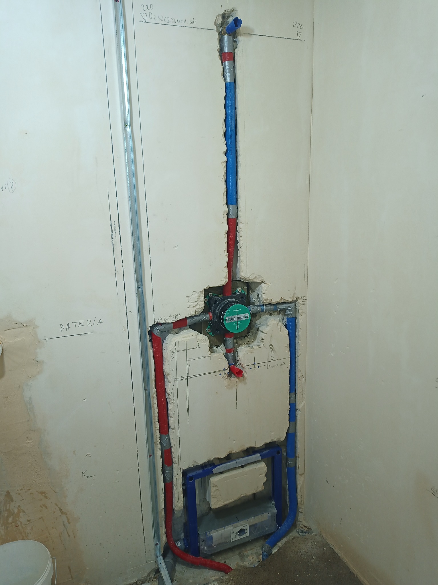 Ściana w trakcie prac instalacyjnych w łazience: widoczne rury (czerwone i niebieskie) prowadzące do zamontowanego podtynkowo zaworu, metalowy profil pionowy, ślady po kuciu w ścianie,...