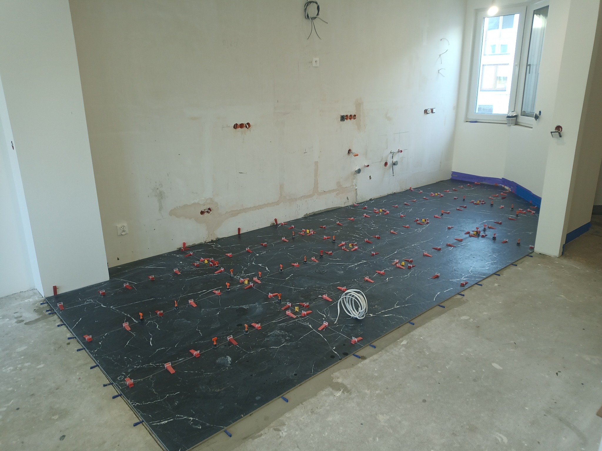 Układanie płytek imitujących marmur na podłodze w łazience, widoczne krzyżyki dystansowe i kliny poziomujące, ściany przygotowane do dalszych prac wykończeniowych, instalacje hydrauliczne...