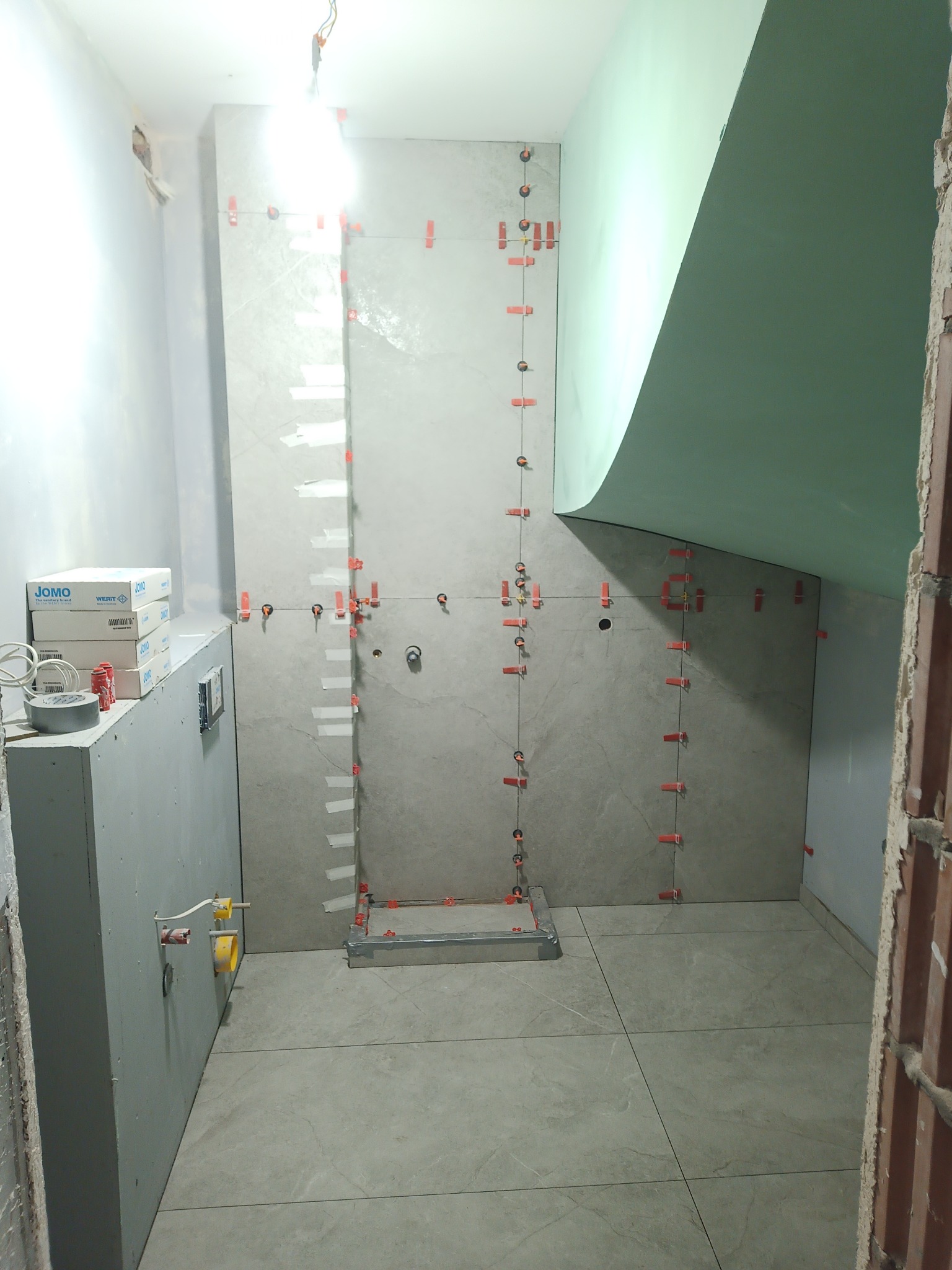 Świeżo wykafelkowana ściana w łazience z użyciem poziomowania płytek, widoczne pudełka z armaturą JOMO i fragment nieotynkowanej ściany.