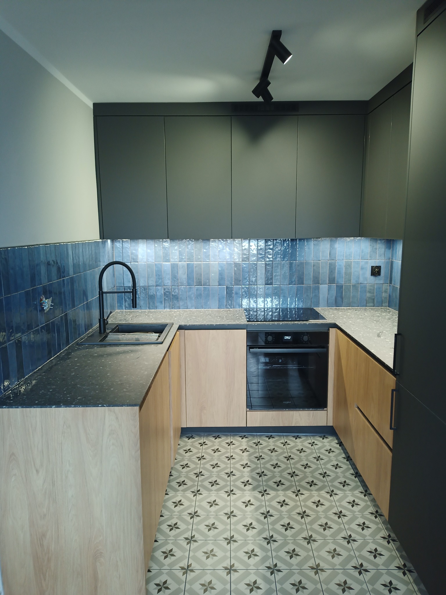 Nowoczesna kuchnia w odcieniach szarości i beżu z niebieskimi płytkami na ścianie, czarną armaturą i wzorzystą podłogą.