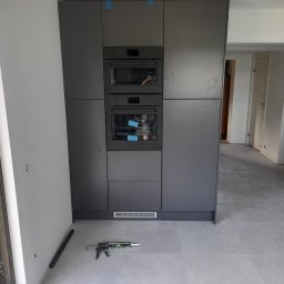 MIDAS Robert Warchoł - Montaż zabudowy kuchennej z wbudowanymi piekarnikami w szarej kolorystyce, widoczny aplikator do silikonu na szarej podłodze z płytek.