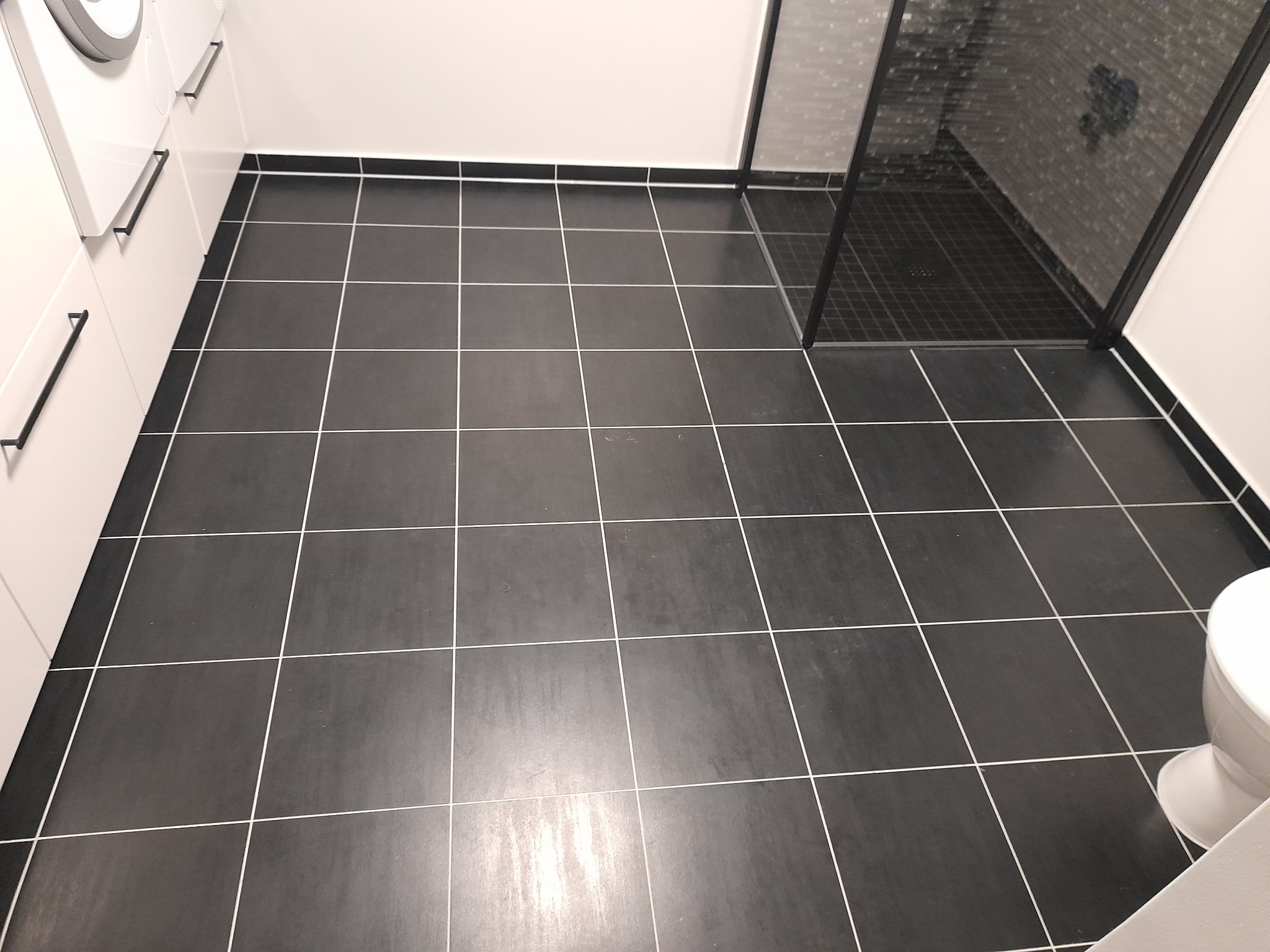 Ciemne, kwadratowe płytki podłogowe z jasnymi fugami w łazience z kabiną prysznicową w Kulno, widok z góry, fragment toalety po prawej.