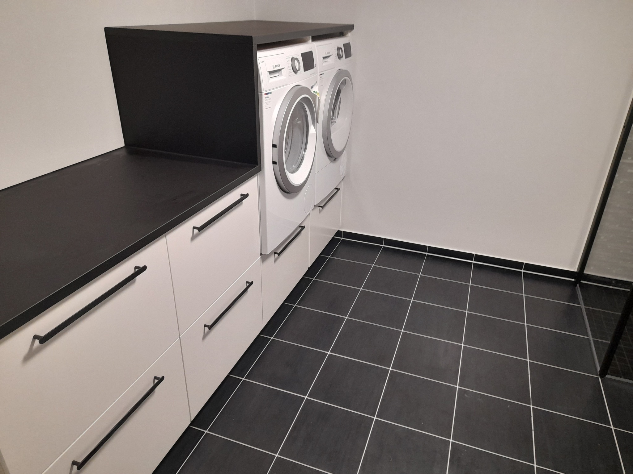 Nowoczesna łazienka z zabudową pralki i suszarki, czarne płytki podłogowe z jasną fugą, białe szafki z długimi, czarnymi uchwytami i czarnym blatem.