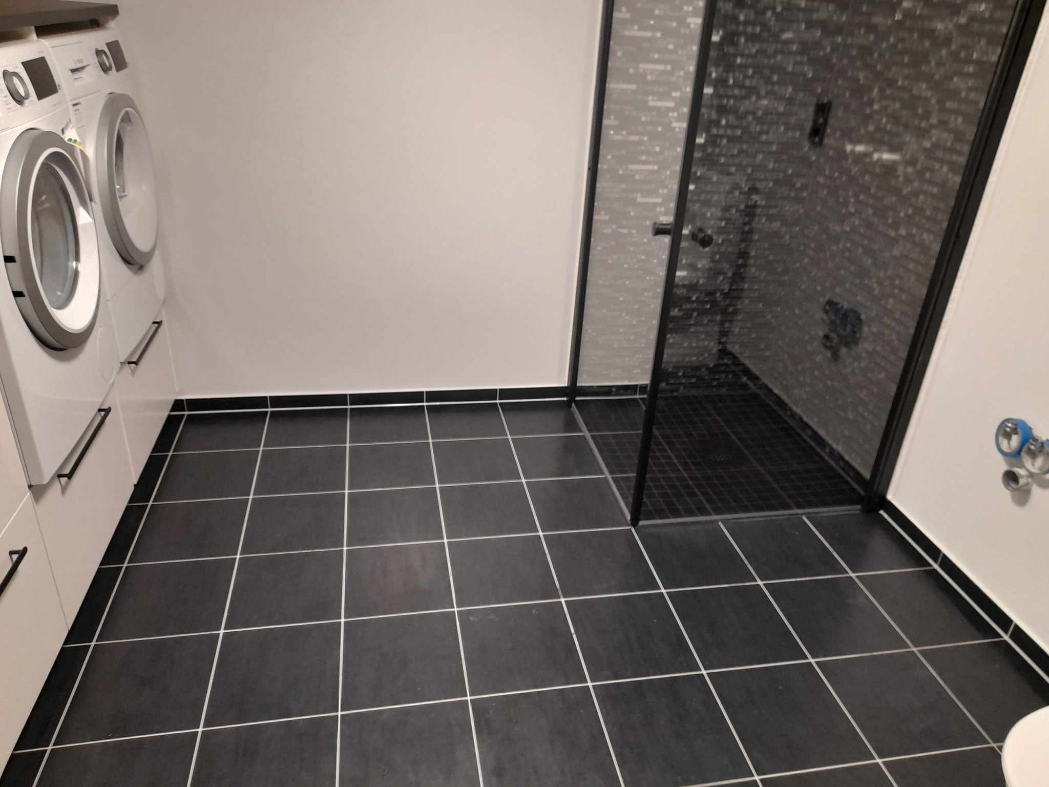 Nowoczesna łazienka z czarnymi płytkami podłogowymi, kabiną prysznicową z czarną ramą i pralką z suszarką w zabudowie.