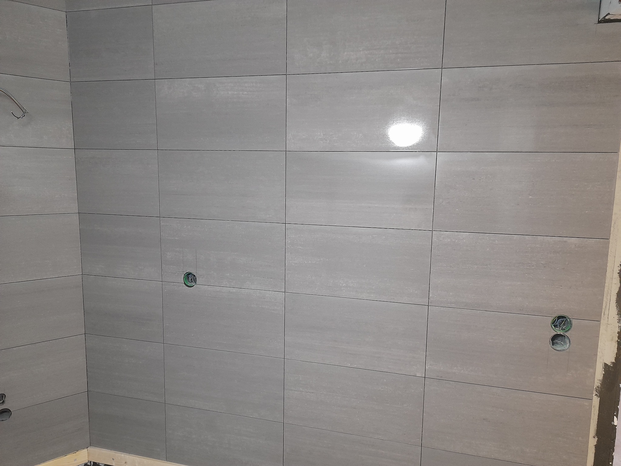 Ściana łazienki wyłożona szarymi, matowymi płytkami ceramicznymi o wyraźnej strukturze, z widocznymi otworami na gniazdka elektryczne i rurę prysznicową, w trakcie prac wykończeniowych.