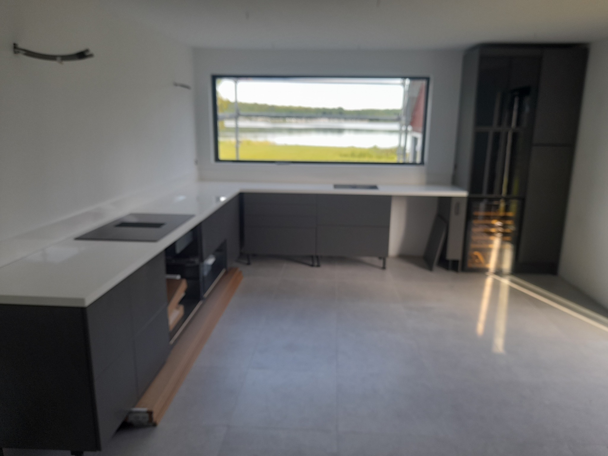 Nowoczesna, szara kuchnia z białym blatem i widokiem na jezioro przez okno. Widoczna zabudowa kuchenna z szufladami i szafkami, oraz wbudowana lodówka na wino. Podłoga wyłożona szarymi płytkami.