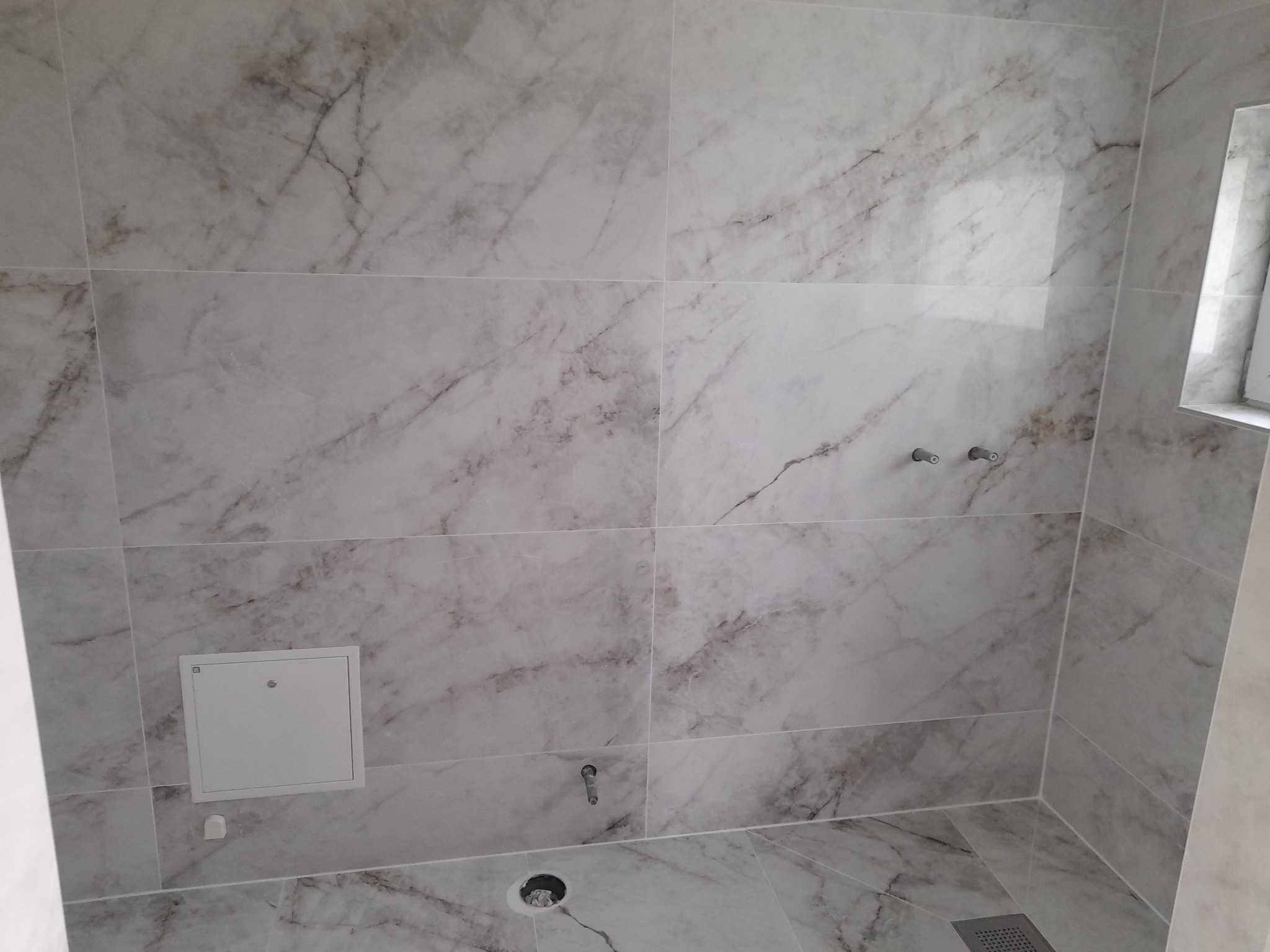 Ściany i podłoga łazienki wyłożone płytkami imitującymi biały marmur z szarymi żyłami, widoczne otwory na armaturę i kratka odpływowa w podłodze, mała klapka rewizyjna na ścianie.