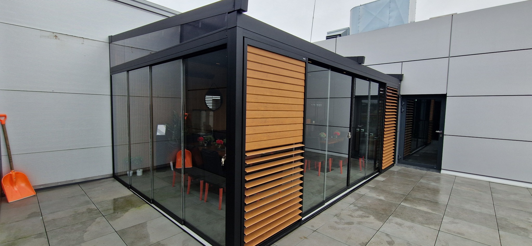Zadaszony taras z przesuwnymi szklanymi ścianami i drewnianymi panelami z żaluzjami, widok z zewnątrz, pomarańczowa łopata oparta o ścianę budynku w tle.