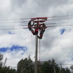Usługi Elektryczne Szymon Kabaciński - Dwóch elektryków w kombinezonach i kaskach na słupie energetycznym, zabezpieczonych linami, wykonuje prace na przewodach wysokiego napięcia na tle pochmurnego nieba.