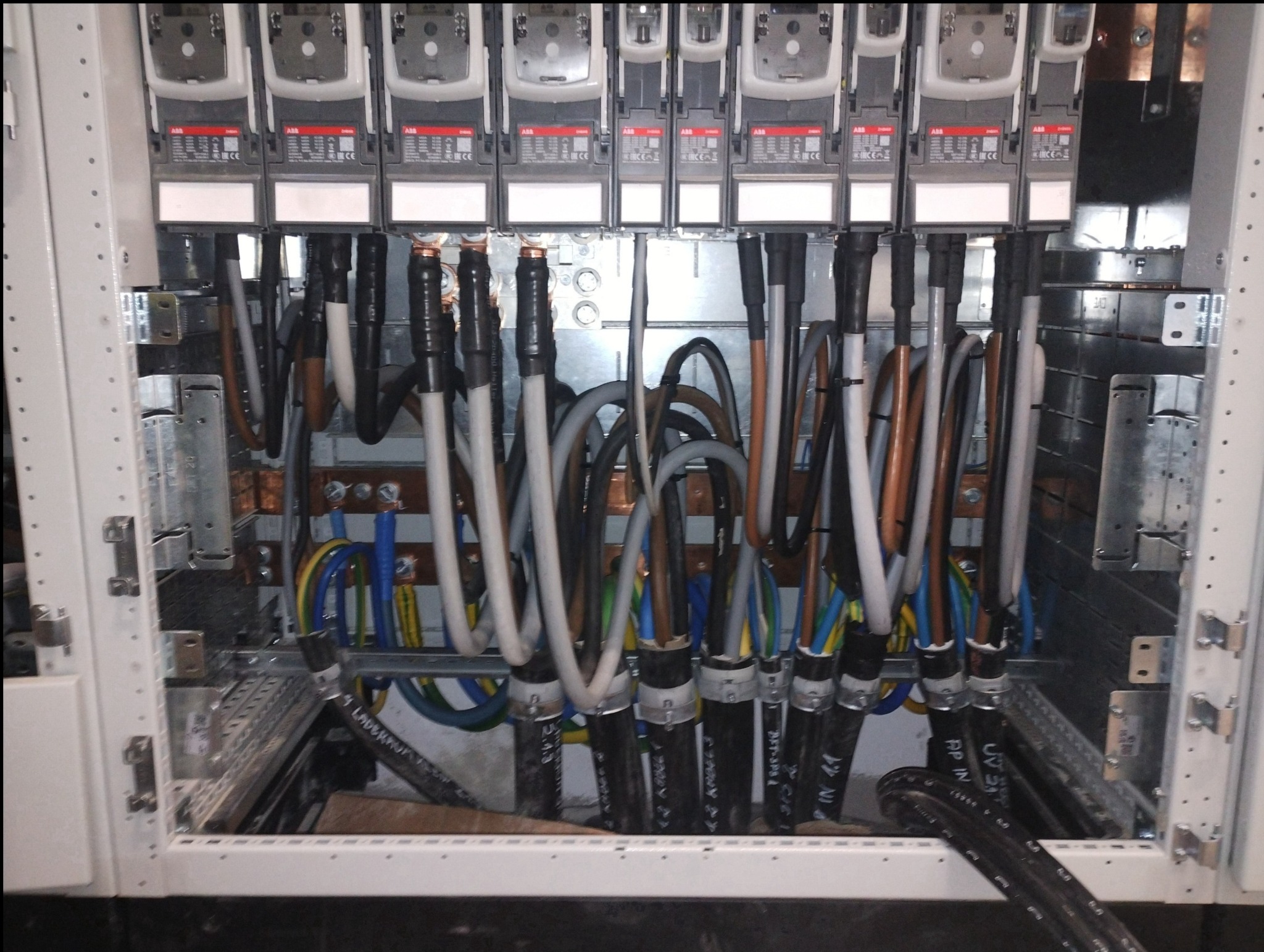 Wnętrze szafy sterowniczej z widocznymi zabezpieczeniami ABB, miedzianymi szynami zbiorczymi i uporządkowanymi przewodami o różnych kolorach, zabezpieczonymi czarnymi osłonami z oznaczeniami.