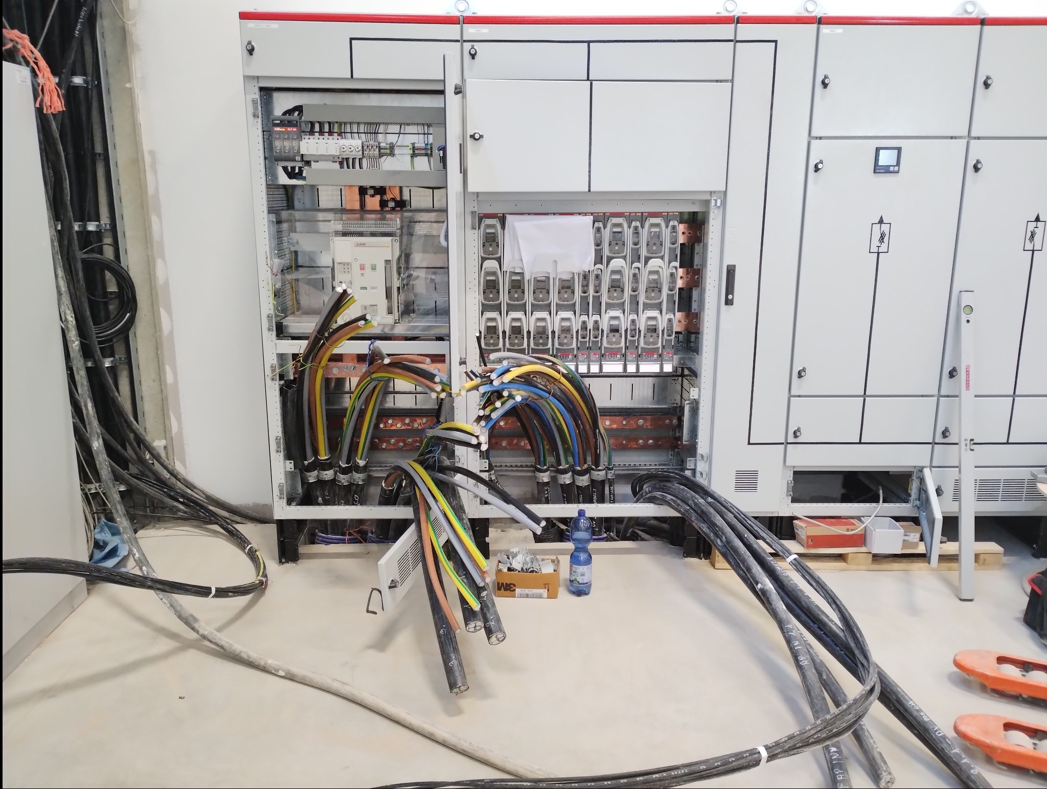 Rozdzielnia elektryczna w trakcie podłączania przewodów, widoczne liczniki, zabezpieczenia oraz kable w różnych kolorach, obok butelka wody i pudełko z akcesoriami.