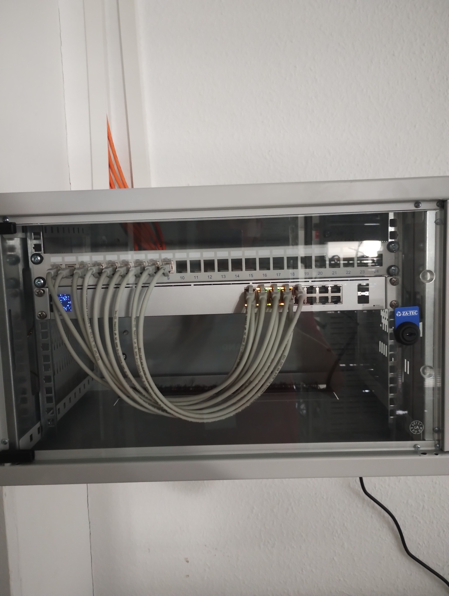 Szafa rack z zamontowanym switchem i patch panelem, połączone szarymi kablami Ethernet. Widoczne diody sygnalizacyjne na switchu oraz oznaczenia portów na patch panelu. Pomarańczowe kable wystają...