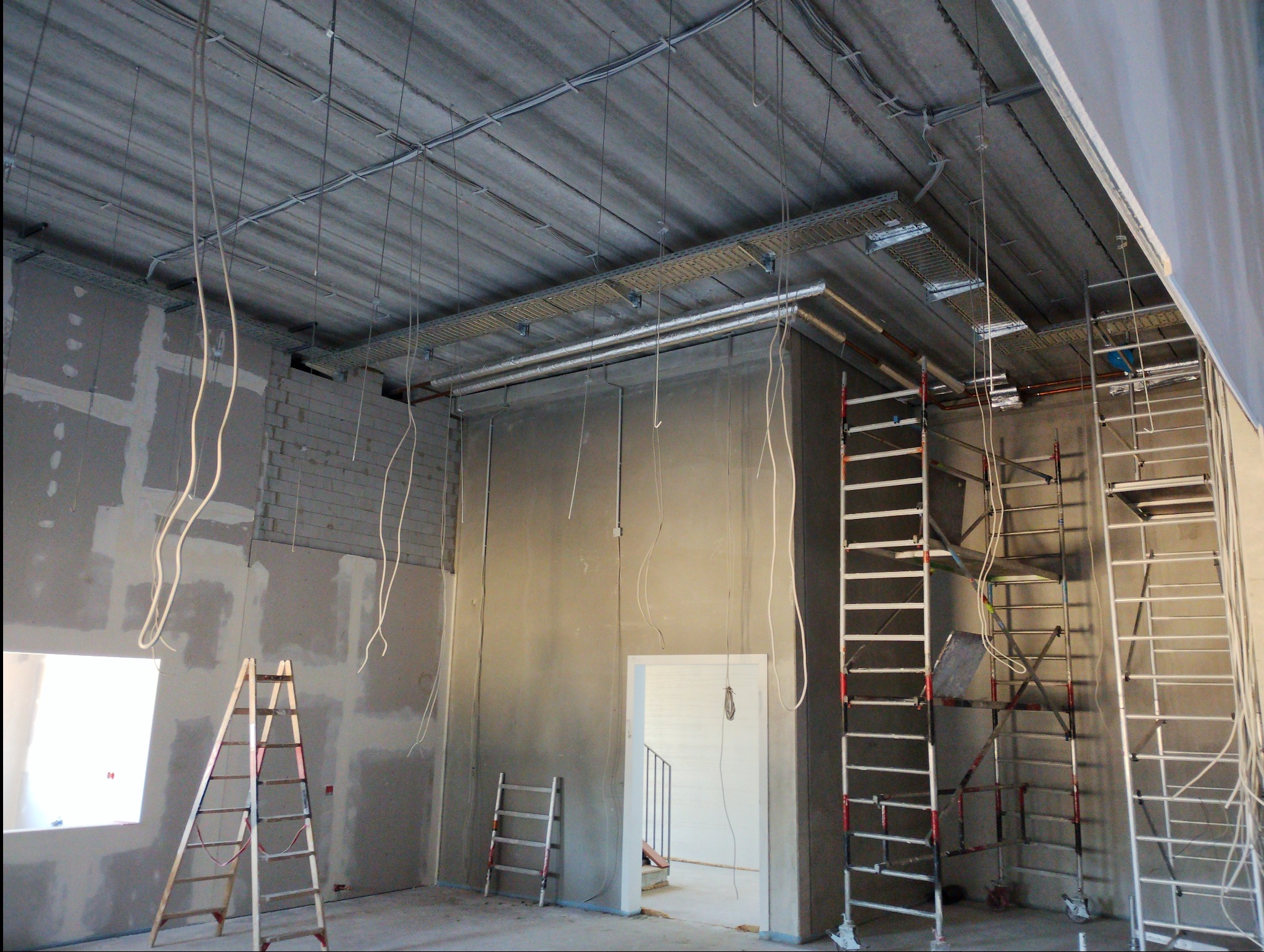 Surowy stan wnętrza w trakcie prac: widoczne kable zwisające z sufitu, koryta kablowe podwieszone do stropu, ściany w stanie surowym, drabiny i rusztowania rozstawione w pomieszczeniu.