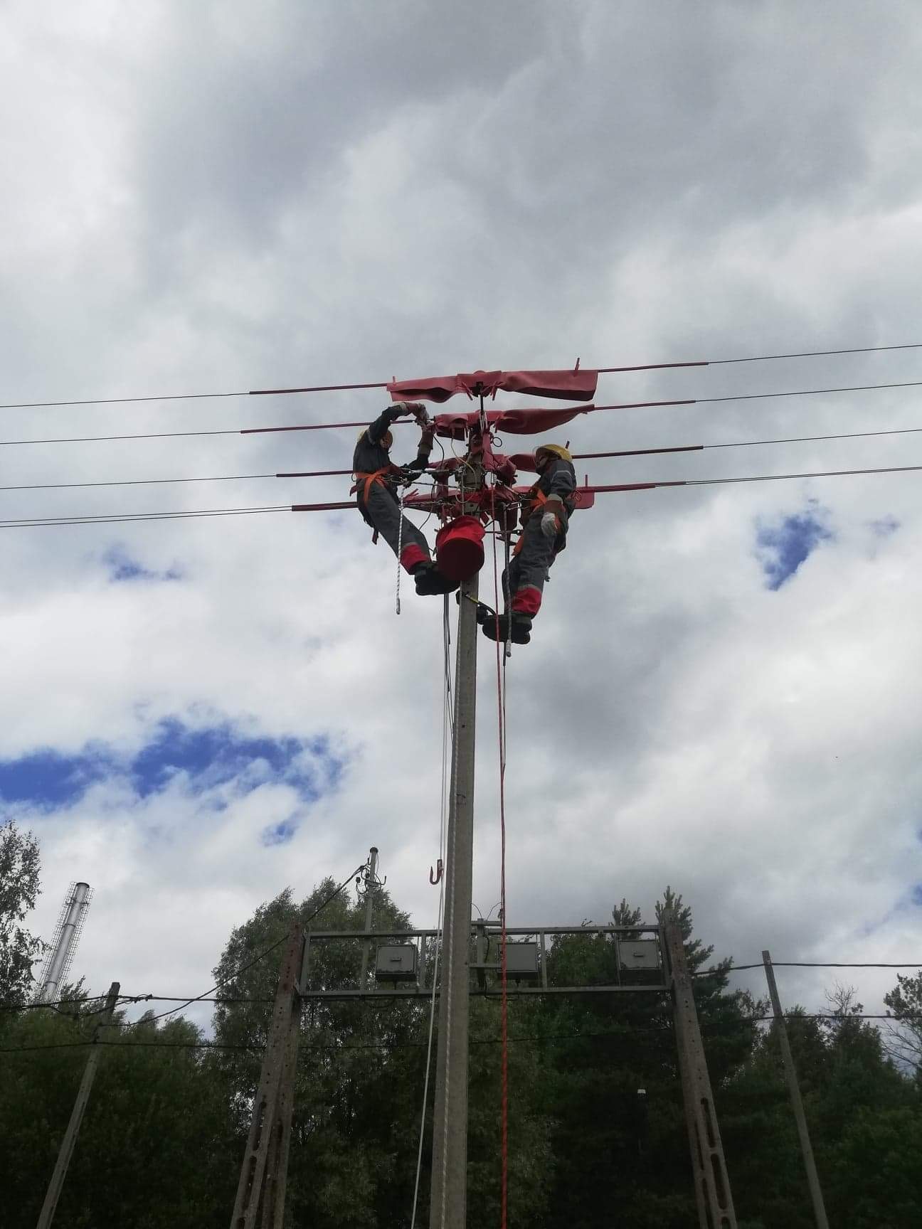Dwóch elektryków w kombinezonach i kaskach na słupie energetycznym, zabezpieczonych linami, wykonuje prace na przewodach wysokiego napięcia na tle pochmurnego nieba.