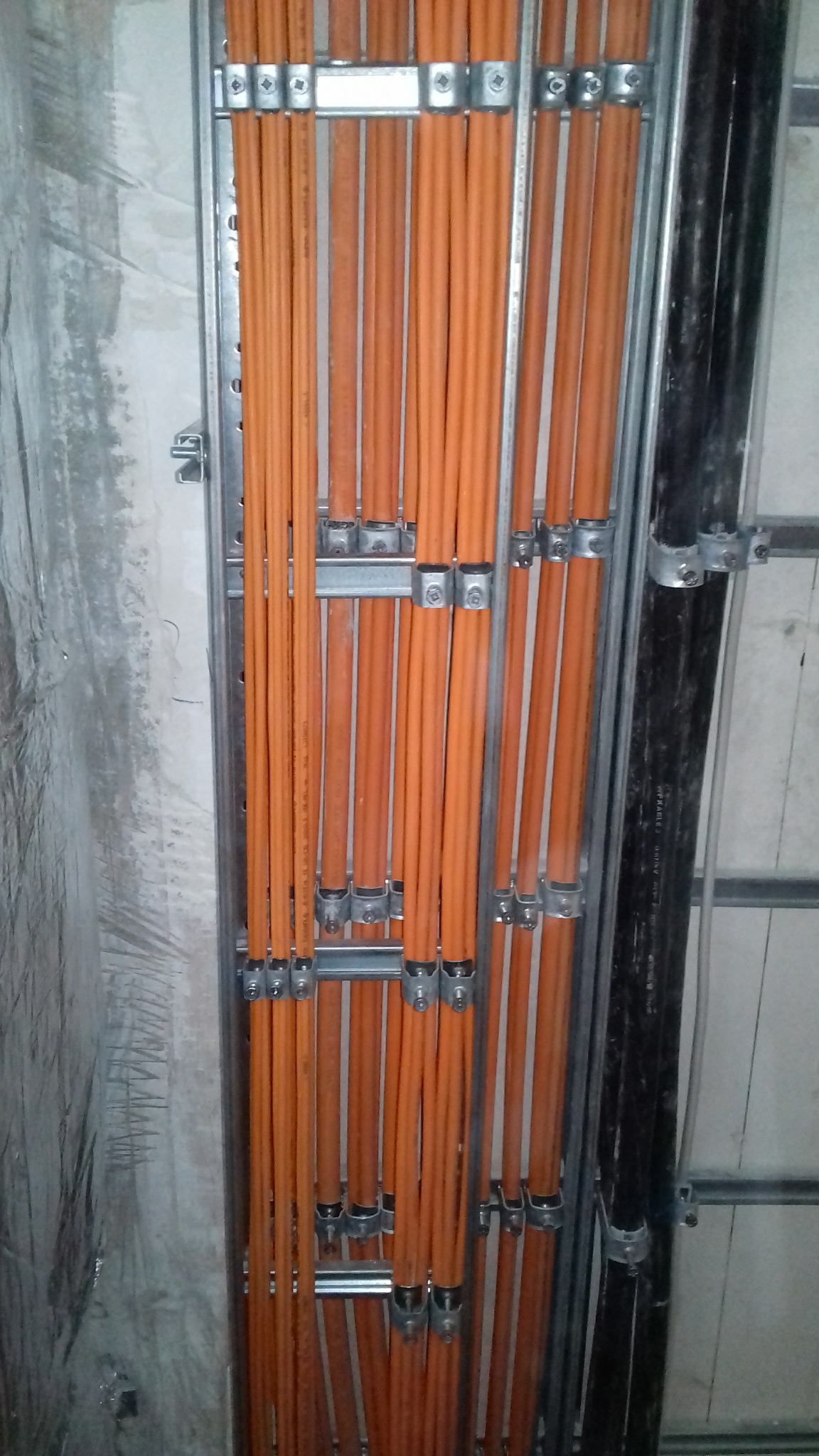 Pomarańczowe i czarne kable elektryczne ułożone pionowo na metalowej konstrukcji montażowej przy ścianie.
