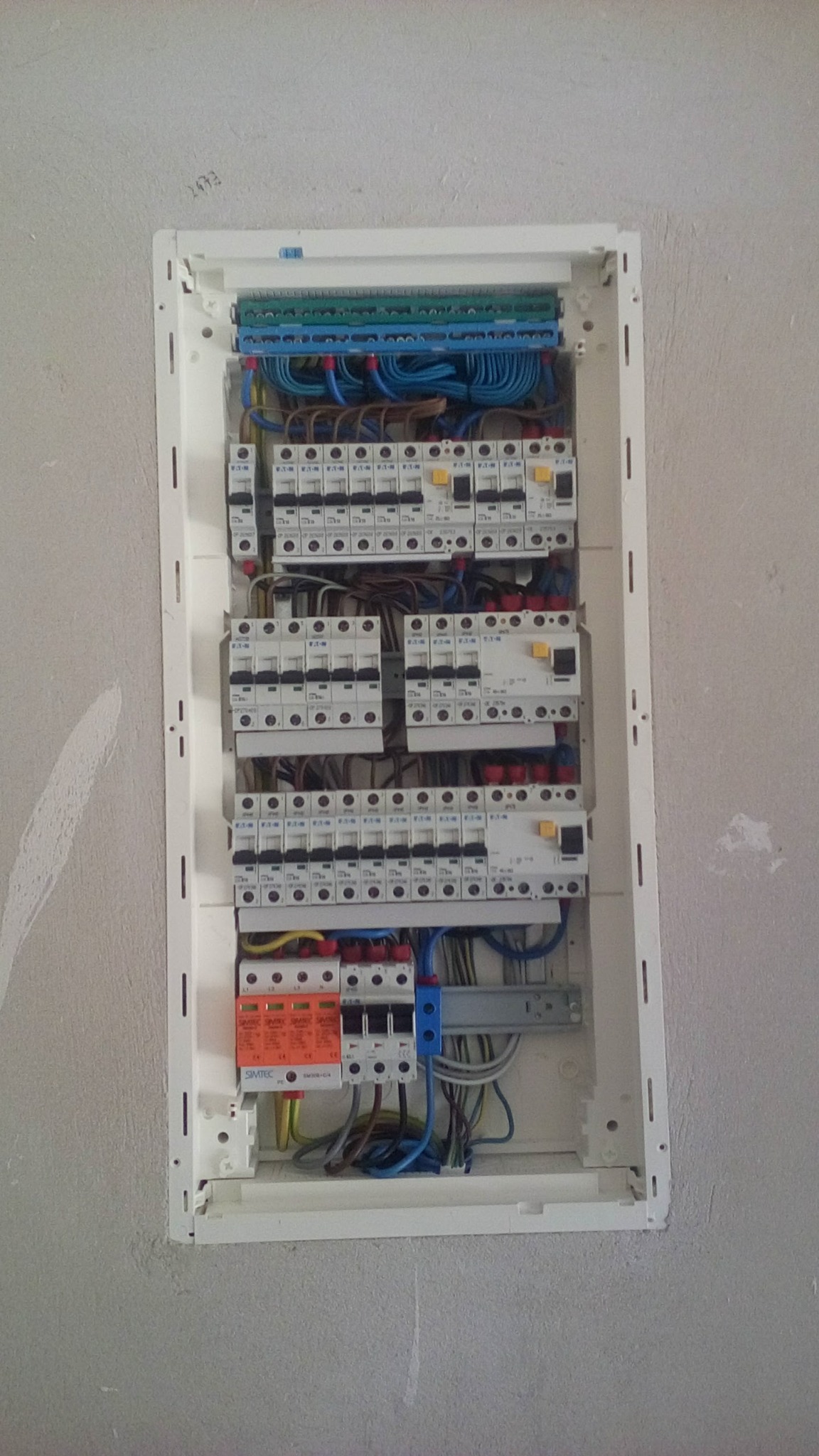 Rozdzielnica elektryczna w białej obudowie, zamontowana w surowej ścianie, widoczne liczne bezpieczniki, kable w różnych kolorach i listwy zaciskowe.