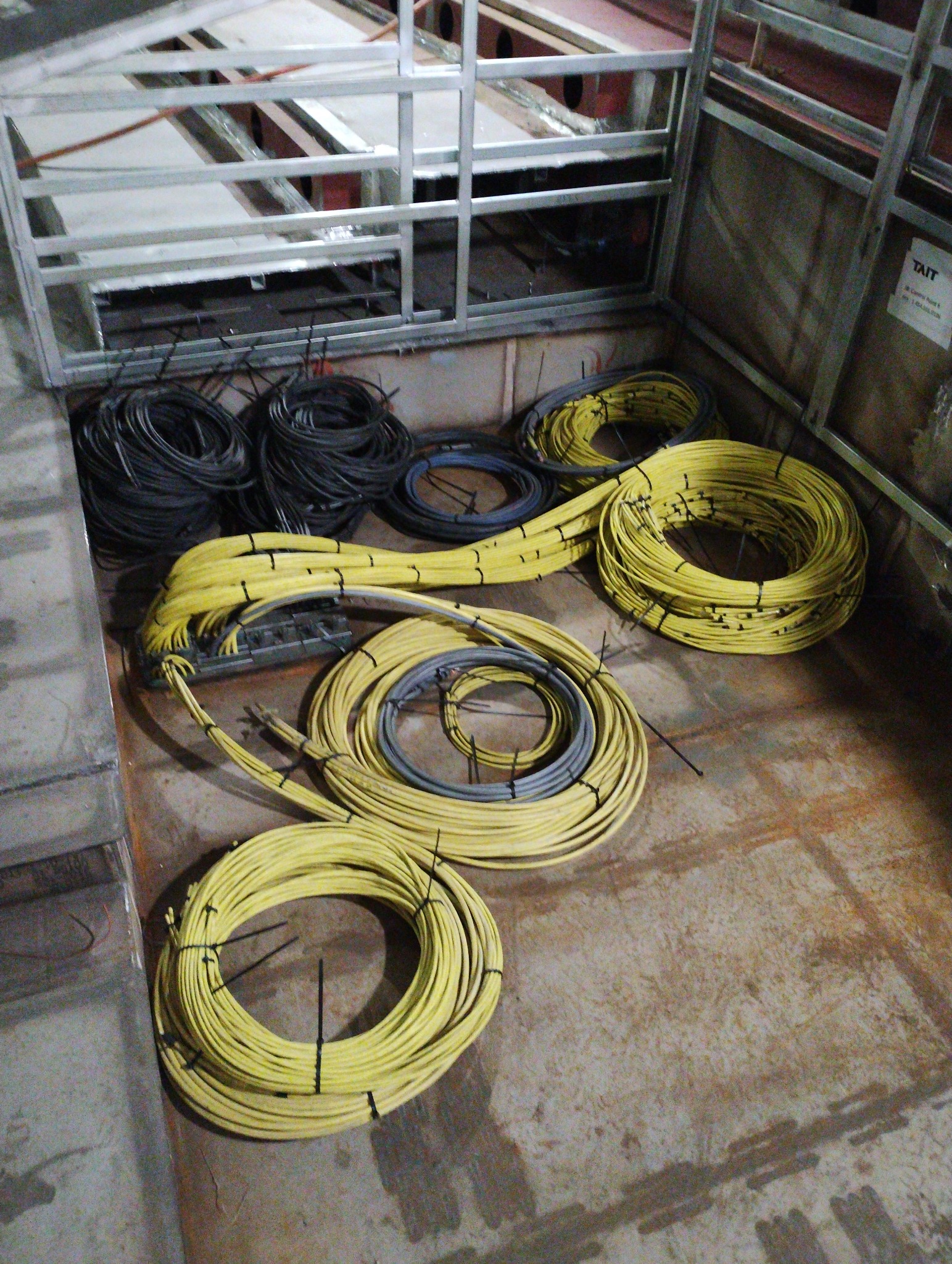Zbiór zwiniętych kabli elektrycznych w kolorze żółtym i czarnym, zabezpieczonych czarnymi opaskami zaciskowymi, leżących na betonowej podłodze w surowym pomieszczeniu z metalową konstrukcją.