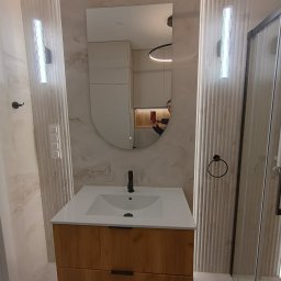 MASTERHOUSE TRL SPÓŁKA Z OGRANICZONĄ ODPOWIEDZIALNOŚCIĄ - Nowoczesna łazienka z drewnianą szafką pod umywalką, podświetlanym lustrem i kabiną prysznicową. Ściany wykończone płytkami imitującymi marmur, oświetlenie LED.