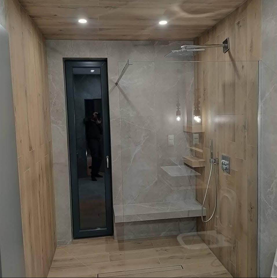 Nowoczesna kabina prysznicowa walk-in z siedziskiem, wykończona płytkami imitującymi drewno i marmur, z widocznym odbiciem osoby w wąskich drzwiach.
