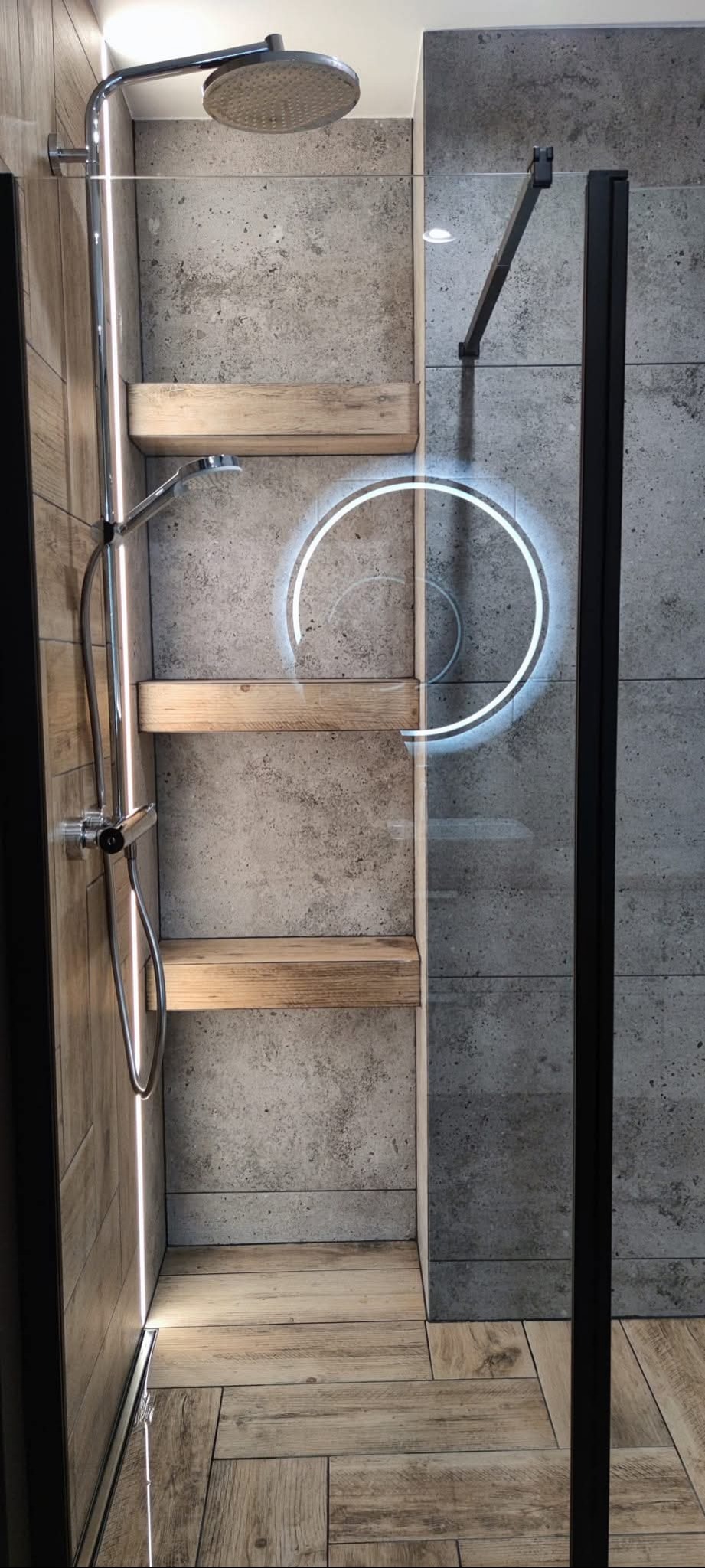 Nowoczesna kabina prysznicowa z drewnianymi półkami i betonowymi płytkami. Okrągłe podświetlenie LED dodaje unikalnego charakteru. Widoczna armatura i szklane drzwi.