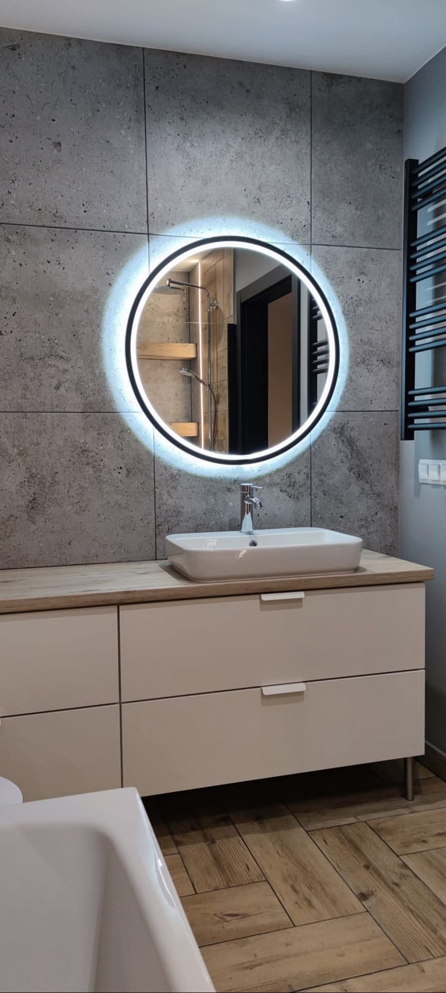 Nowoczesna łazienka z okrągłym lustrem LED, umywalką nablatową i szarą płytką imitującą beton. Widoczny fragment wanny i drewnopodobna podłoga.